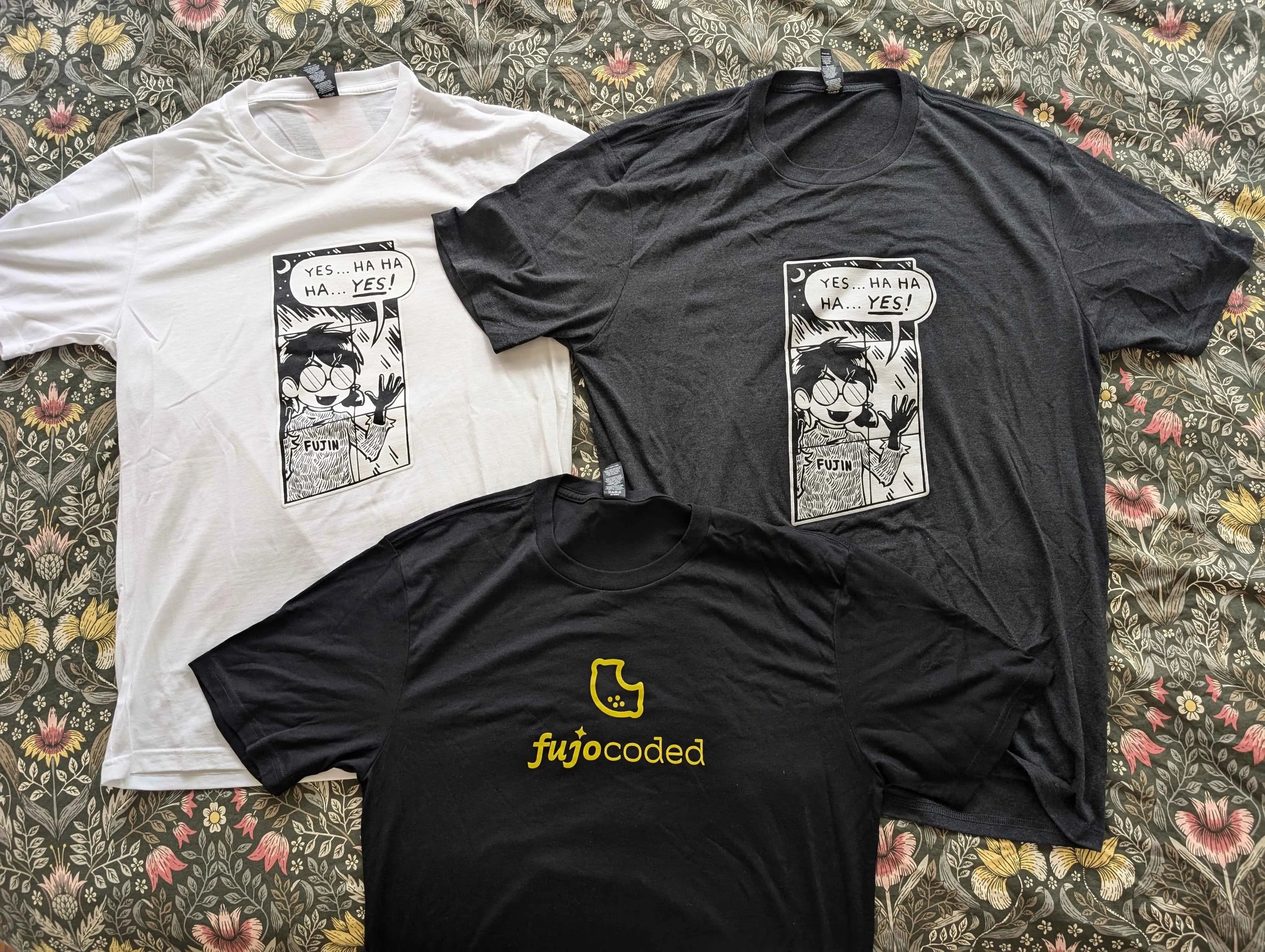 Photograph of three shirt options: Fujin Sickos meme parody as a black T-shirt and a white T-shirt, as well as the FujoCoded lemon logo on a black T-shirt.